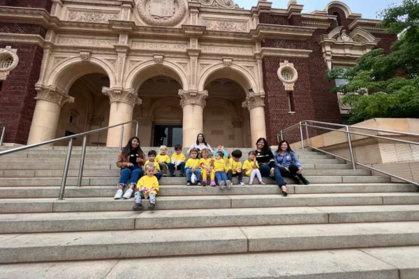 Group of children and adults sitting on steps in front of a grand historic building.